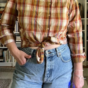 May include: A plaid button-up shirt in shades of brown, orange, and white is tied at the waist. The shirt has a rounded collar and long sleeves. The shirt is paired with light blue denim shorts.