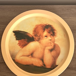 May include: A round, peach-colored tray featuring a classical painting of a cherub with red hair and wings. The cherub is resting its chin on its hand, with a thoughtful expression. The tray has a peach-colored rim.