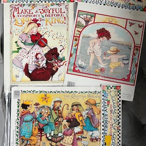 May include: Several Mary Engelbreit iron-on transfers are displayed. One features a girl playing a violin with a dog, another shows children at the beach, and a third depicts a Christmas pageant scene. The transfers have colorful illustrations and text.