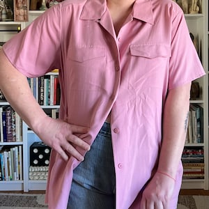 May include: A light pink button-down shirt with short sleeves and two chest pockets. The shirt is unbuttoned at the waist, revealing blue jeans. The shirt has a classic collar and a relaxed fit, suitable for casual wear.