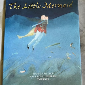 May include: A hardcover book titled "The Little Mermaid" with gold lettering. The cover features an illustration of a person in a red coat and white pants, with a mermaid tail, swimming underwater. The author's names are also printed on the cover.