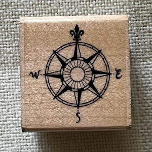 May include: A square wooden rubber stamp featuring a black ink illustration of a compass rose. The design includes a fleur-de-lis at the north point and the letters W, E, and S marking the west, east, and south directions on the circular frame.