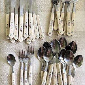 May include: A collection of stainless steel flatware featuring off-white handles with delicate floral patterns in pink and blue. The set includes various knives, forks, and spoons, all displaying a consistent design with metallic heads and decorative handles.