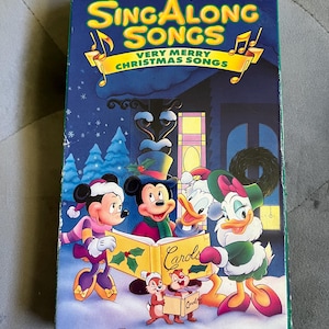May include: Disney's Sing Along Songs: Very Merry Christmas Songs VHS tape. The cover features Mickey Mouse, Minnie Mouse, Donald Duck, and Daisy Duck singing carols in a winter scene. The title is in yellow and the volume is eight.