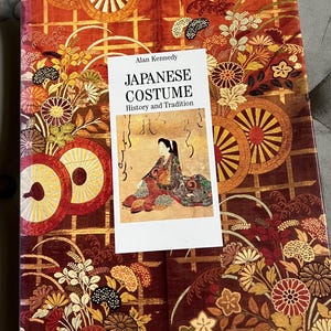 May include: A book titled "Japanese Costume History and Tradition" by Alan Kennedy. The book cover features a detailed floral and geometric pattern in shades of red, gold, and brown. A smaller image of a person in traditional Japanese attire is on the cover.