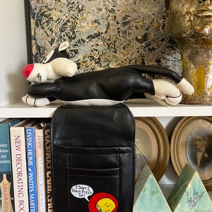 May include: A black leather bag with a zippered pocket and a Tweety Bird patch that says "I Tawt I Taw a Puddy Tat!" The bag is sitting on a shelf with books and a black and white cartoon cat plush toy.