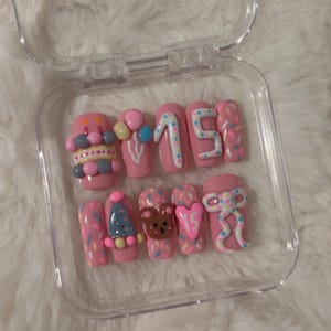 May include: A set of pink press-on nails with various designs, including a cupcake, balloons, the number 15, a Christmas tree, a teddy bear, and a bow. The nails are decorated with sprinkles and other embellishments, and are displayed in a clear plastic case.