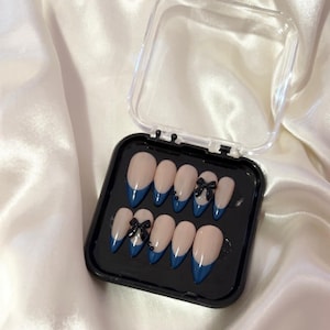 May include: A set of artificial nails in a black case. The nails have a nude base with a dark blue French tip and black bow accents. The case is clear and open, set against a white satin background.