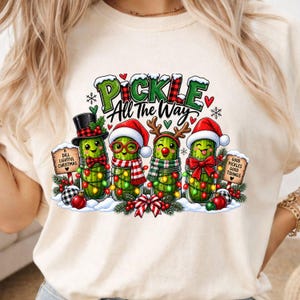 Könnte beinhalten: Ein cremefarbenes T-Shirt mit einer Grafik von vier in Gläsern steckenden, weihnachtlich gekleideten Gewürzgurken. Der Text lautet "PICKLE All the Way", "A DILL LIGHTFUL CHRISTMAS" und "GOOD PICKLES GOOD TIDINGS". Die Gurken tragen Mützen und Schals.