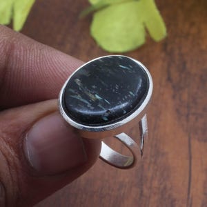 May include: A silver ring featuring a large, oval-shaped black stone with subtle grey and blue inclusions. The stone is set in a smooth, polished silver bezel, and the band consists of a split-shank design that tapers toward the back of the ring.
