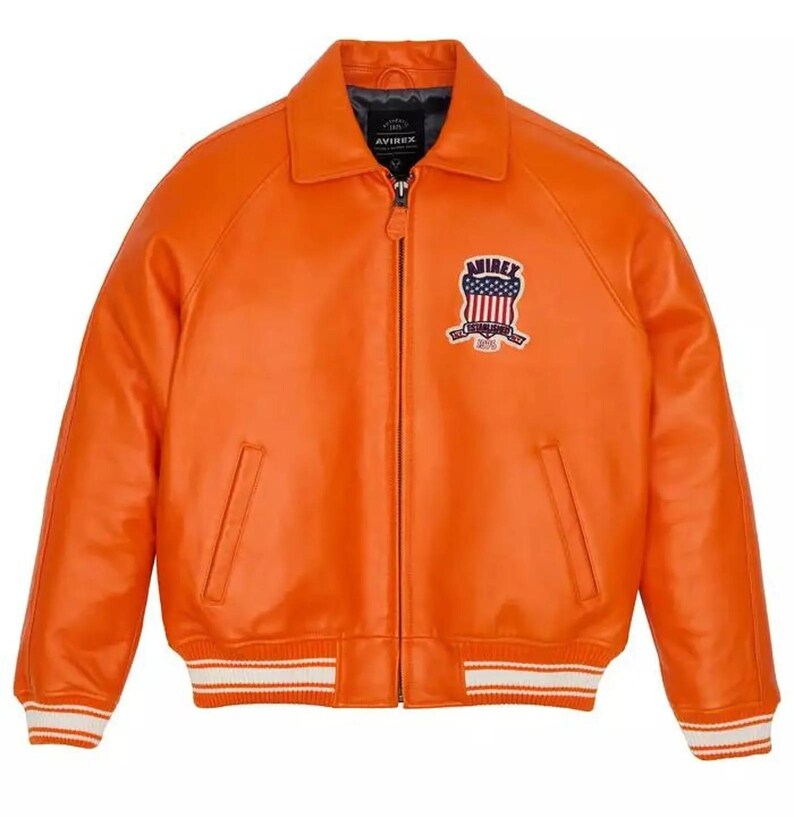 May include: Bright orange leather jacket with a zip closure, collar, and two front pockets. The jacket has white and red striped trim on the cuffs and hem. A chest patch displays the text "AVIREX" and a patriotic emblem.