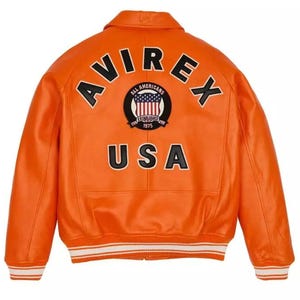 May include: Bright orange leather jacket with the word "AVIREX" arched across the top and "USA" at the bottom. A circular patch in the centre features the words "ALL AMERICANS" and "ESTABLISHED 1975" around a red, white, and blue emblem. The jacket has white and black striped cuffs.