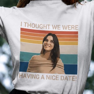 May include: A white long-sleeve t-shirt featuring a graphic print of a person with long dark hair wearing a beige off-the-shoulder top. The background of the graphic has horizontal stripes in red, orange, yellow, teal, and blue with the text I THOUGHT WE WERE HAVING A NICE DATE!