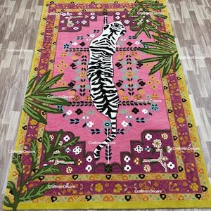 May include: A rectangular area rug featuring a central white tiger with black stripes on a vibrant pink background. The rug is decorated with green palm leaves, small white and yellow floral patterns, and a wide border in shades of purple and lime green.