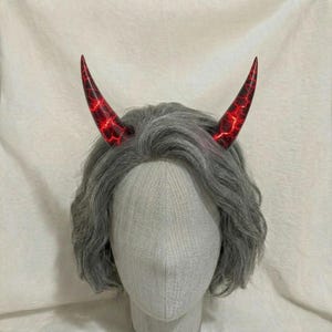 May include: Red and black devil horns with a crackle pattern, set on a grey wig. The horns are pointed and have a glowing red interior. The wig features wavy, grey hair.
