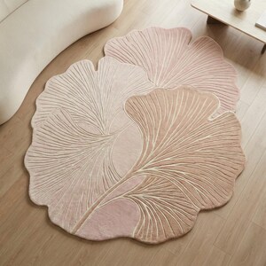Könnte beinhalten: Drei blattförmige Teppiche in verschiedenen Rosa- und Beigetönen. Die Teppiche haben ein detailliertes, strukturiertes Design, das die Adern eines Blattes nachahmt. Sie sind auf einem hellen Holzboden angeordnet und schaffen eine weiche, natürliche Ästhetik.