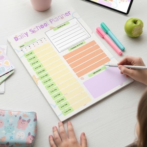 May include: A Daily School Planner with a pastel color scheme. The planner features a schedule from 6:00 AM to 6:00 PM, with sections for study plans and notes. A person is writing on the planner with a silver pen. Other items include pens, stickers, and a pencil case.