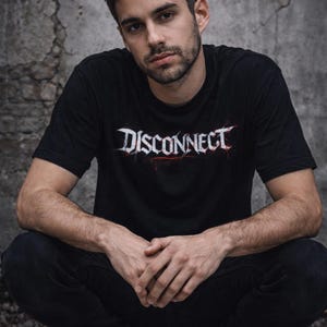 Puede incluir: Una persona con una camiseta negra con la palabra "DISCONNECT" en letras blancas y rojas. La persona lleva pantalones negros y zapatos negros. El fondo es una pared gris texturizada.