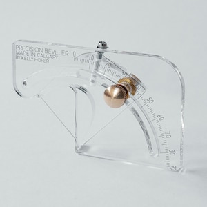 Può includere: Un bisellatore di precisione trasparente con una scala curva da 0 a 90 gradi. Il testo "PRECISION BEVELER MADE IN CALGARY BY KELLY HOFER" è stampato in alto a sinistra. Una manopola in ottone regola l'angolo.