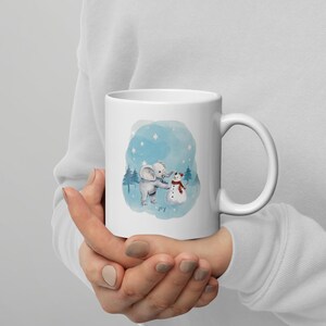 Pode incluir: Caneca de cer&acirc;mica branca com uma ilustra&ccedil;&atilde;o em aquarela de um elefante cinzento e um boneco de neve. O design inclui um fundo azul com estrelas e pinheiros, e a palavra "joy" na parte inferior.