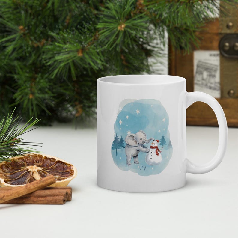 Pode incluir: Caneca de cer&acirc;mica branca com uma ilustra&ccedil;&atilde;o de inverno de um elefante e um boneco de neve. O design apresenta um fundo azul em aquarela, flocos de neve e a palavra "joy".