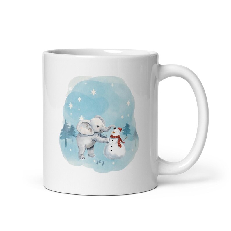 Pode incluir: Caneca de cer&acirc;mica branca com uma ilustra&ccedil;&atilde;o em aquarela de um elefante cinza e um boneco de neve. O elefante est&aacute; tocando o cachecol do boneco de neve. Um fundo azul em aquarela com estrelas e pinheiros completa o design. A palavra "joy" est&aacute; escrita em it&aacute;lico.