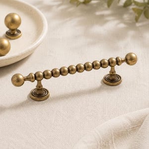 Peut inclure: Ensemble de quincaillerie de meuble en laiton antique. L'ensemble comprend une poignée à perles et deux boutons ronds. La poignée comporte une série de petites sphères et est montée sur une surface crème. Les boutons sont sur une assiette blanche.