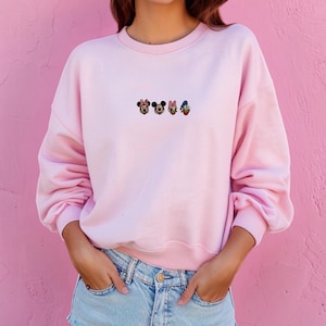 May include: A light pink crewneck sweatshirt featuring a small, centred embroidery of four classic cartoon character heads in a horizontal row, including Minnie Mouse, Mickey Mouse, Daisy Duck, and Donald Duck, paired with light blue denim shorts.
