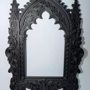 May include: A black gothic-style picture frame with elaborate carvings. The frame has an arched top with detailed floral and geometric patterns. The design incorporates columns and decorative elements, giving it a vintage appearance. The frame is made of a dark material.