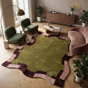 May include: An area rug with an irregular, organic shape featuring a large olive green centre. The border consists of a geometric, stepped pattern in alternating blocks of deep burgundy and soft dusty pink, creating a distinct contrast.