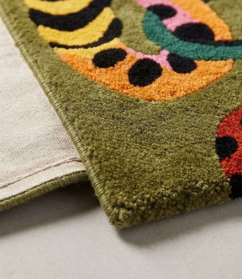 Peut inclure: Un tapis tuft&eacute; pr&eacute;sentant une base vert olive profond avec un motif vibrant de formes jaunes, orange, roses et rouges accentu&eacute;es par des taches noires. Le tapis a un poil &eacute;pais et textur&eacute; avec un bord fini et un envers en tissu beige.