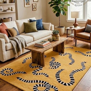May include: A rectangular area rug featuring a vibrant yellow background with a repeating pattern of black and white striped snakes. The rug is positioned on a wooden floor in a living room setting. A circular logo in the corner reads Indian Luxury Rugs Hand Made Est 1978 Rugsin.