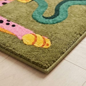 Peut inclure: Un tapis touffet&eacute; &agrave; fond vert olive pr&eacute;sentant un membre animal &agrave; motifs roses et orange avec des taches noires et une patte jaune, ainsi qu'une forme sinueuse vert fonc&eacute; rappelant un serpent avec une finition en relief textur&eacute;e.