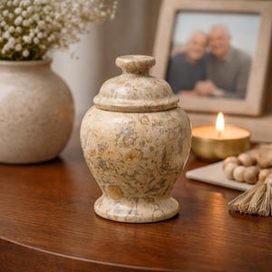 以下が含まれることがあります： 木製の表面に置かれた、斑点模様のベージュ色の蓋付き骨壺。 骨壺は丸みを帯びた本体と、蓋に小さな円形のつまみがあります。 白い花の入った花瓶と額入りの写真が背景にあります。