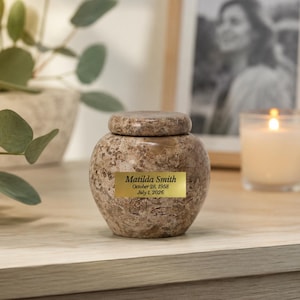 May include: A round, brown, mottled stone urn with a matching lid. A rectangular gold-coloured plaque is attached to the front, featuring the black engraved text: Matilda Smith, October 28, 1958, July 1, 2026. The urn has a smooth, polished finish.