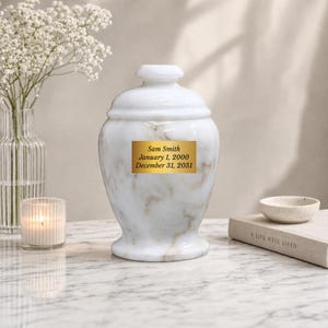 May include: A white marble-patterned cremation urn with a matching lid and a rectangular gold plaque on the front. The plaque features the engraved text: Sam Smith, January 1, 2000, December 31, 2031. The urn has a smooth, rounded silhouette.