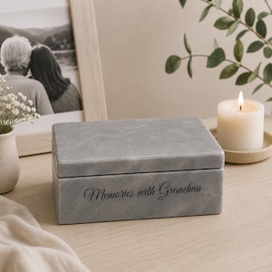 May include: A rectangular grey stone keepsake box with a smooth finish and a lid. The front of the box features the engraved black text "Memories with Grandma" in a cursive script font. The box is positioned on a flat surface.
