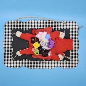 May include: A whimsical Santa Claus figure made of red fabric, adorned with various colorful buttons and embellishments. The figure is set against a black background, framed by a black and white checkered border, and suspended by a jute string.