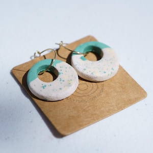 Peut inclure: Une paire de boucles d'oreilles circulaires faites à la main. Chaque boucle d'oreille est en forme de donut, avec une moitié supérieure turquoise et une moitié inférieure blanc cassé mouchetée. Les boucles d'oreilles sont sur des crochets argentés.