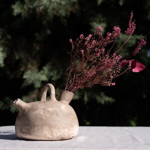Puede incluir: Un recipiente de cerámica de color beige claro con forma redondeada, un pequeño pico y un asa central, que contiene un ramo de flores de brezo secas de color rosa intenso. La superficie de arcilla tiene una textura natural y mate.