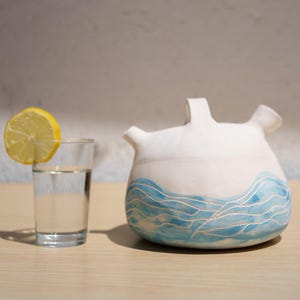 Puede incluir: Un recipiente de cerámica blanca con cuerpo redondeado y dos boquillas, que presenta un patrón de olas azules pintado a mano en la base. Junto a él hay un vaso transparente lleno de agua, decorado con una rodaja de limón amarillo en el borde.