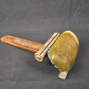 Può includere: Un antico strumento da cucito con manico in legno e meccanismo in metallo. Presenta un cuscino in velluto verde e un rullo in metallo. Il manico in legno è rettangolare con una striscia di metallo. Probabilmente per cucire o creare.