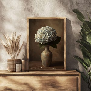 Può includere: Stampa incorniciata raffigurante una natura morta di un bouquet di ortensie blu in un vaso decorativo. Il vaso è posto su una superficie di legno, con uno sfondo neutro. La stampa è in una cornice di legno.