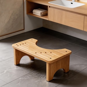 Peut inclure: Un tabouret de salle de bain en bambou brun clair avec une découpe incurvée et un dessus fendu. Le tabouret a quatre pieds et est conçu pour une utilisation dans une salle de bain. Le tabouret est en bambou naturel.