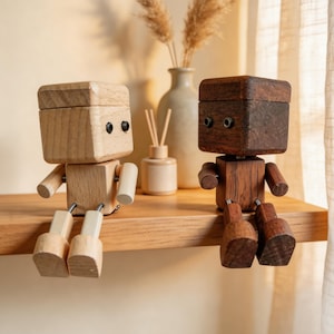 Może przedstawiać: Dwie drewniane figurki robotów, jedna jasna i jedna ciemna, siedzą na drewnianej półce. Roboty mają głowy i ciała w kształcie klocków z ruchomymi ramionami i nogami. Jasny robot ma naturalne drewniane wykończenie, a drugi jest ciemnobrązowy.