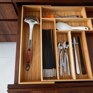 May include: A wooden drawer organizer filled with kitchen utensils. The drawer contains a slotted ladle, black chopsticks, silverware, a rice paddle, and a silicone brush. The organizer is made of light-colored wood.