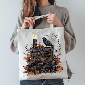 May include: A white canvas tote bag featuring a graphic print of a stack of five dark, weathered books topped with a lit black candle. A black crow sits on the top book, surrounded by brown autumn leaves, bare branches, a spider web, and a spider.