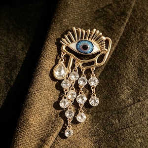 Könnte beinhalten: Goldfarbene Augenbrosche mit blauer Iris und weißem Hintergrund. Die Brosche hat einen tropfenförmigen Edelstein und mehrere hängende, klare Kristalle. Das Augendesign ist auf einem dunkelgrünen Stoff angebracht.