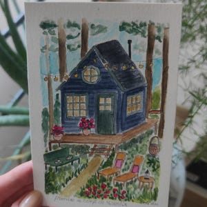 Può includere: Un dipinto ad acquerello su carta che raffigura una capanna blu scuro immersa in un bosco. La capanna presenta una finestra rotonda, una terrazza in legno con fiori rosa in vaso e luci a corda. Sotto, un giardino include una panchina verde, due sedie a sdraio e fiori rossi, con il testo francese scritto a mano "prendre le temps de ralentir" in basso.
