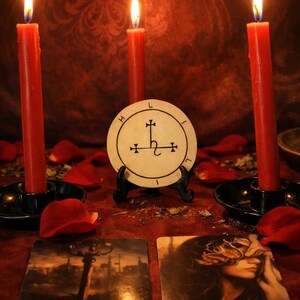 May include: A mystical scene featuring three red candles with lit flames, a circular emblem, and two tarot cards. The cards read "VERTIGO" and "EPIPHANY". Scattered rose petals add to the ambiance. The candles are in black holders.